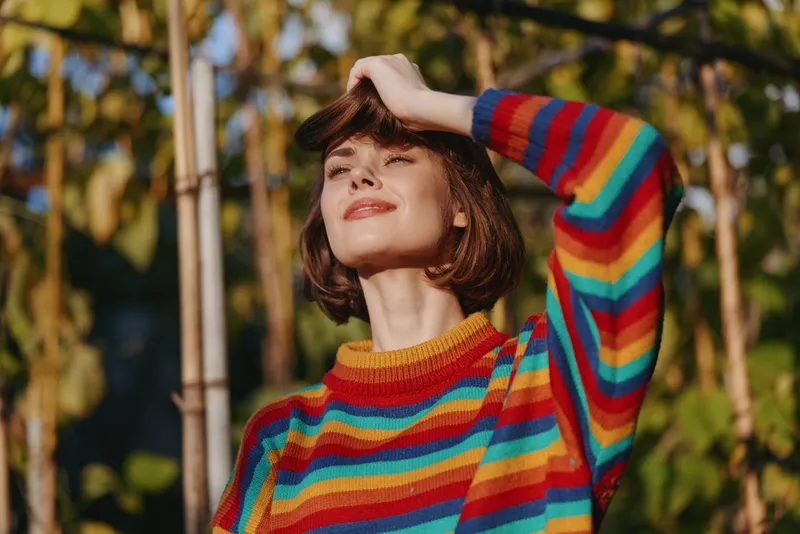 Young woman in her 20s wearing a rainbow sweater and short hair, smiling outdoors in warm sunlight, hand on forehead in a tropical garden portrait for lifestyle and fashion vibe.