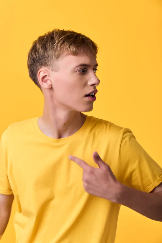 Young man in a bright yellow shirt with a pointing gesture against a matching yellow studio backdrop, conveying bold fashion style and confident direction