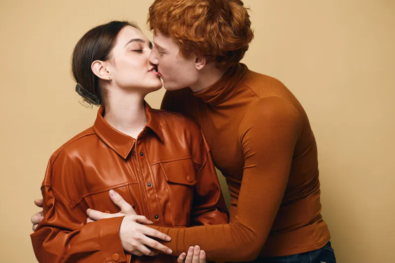 Young couple kissing and embracing each other in casual clothing against beige background. Man with red hair and woman in leather jacket sharing intimate moment of affection.