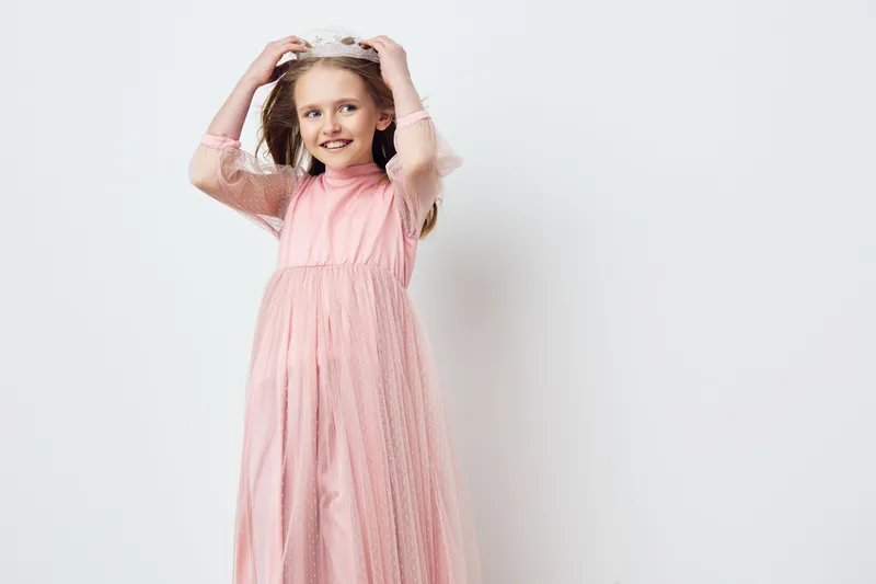 Cute girl in a pink dress with a crown, smiling joyfully against a soft white background, ideal for children s fashion and celebration themes