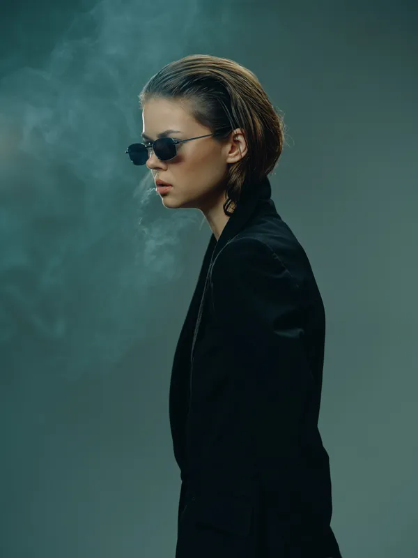Stylish woman in sunglasses with slicked-back hair exhaling smoke against dark background, wearing black jacket, moody atmosphere.