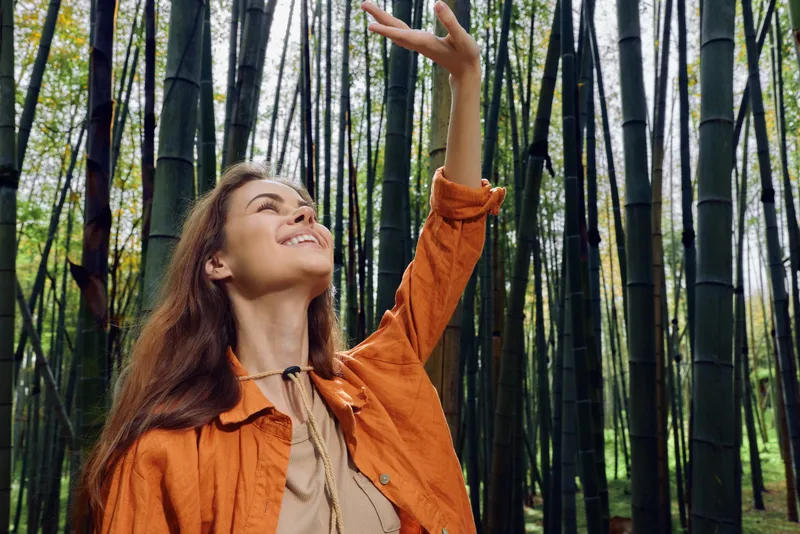 Woman in bamboo forest smiles and reaches upward in a nature portrait, enjoying sunlight and serenity; long hair and orange jacket convey joy and peaceful outdoor happiness.