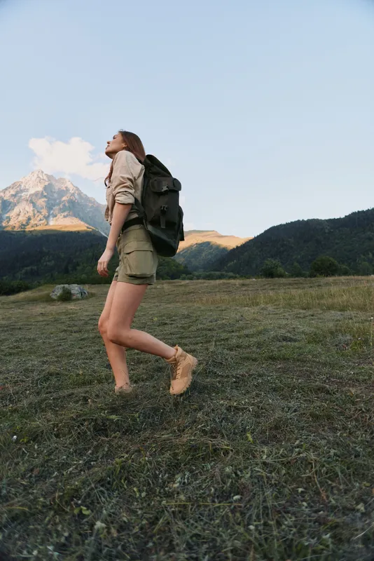 A young woman hikes through a grassy valley with a backpack, wearing shorts and boots, gazing toward distant mountains. A candid outdoor lifestyle moment in nature.
