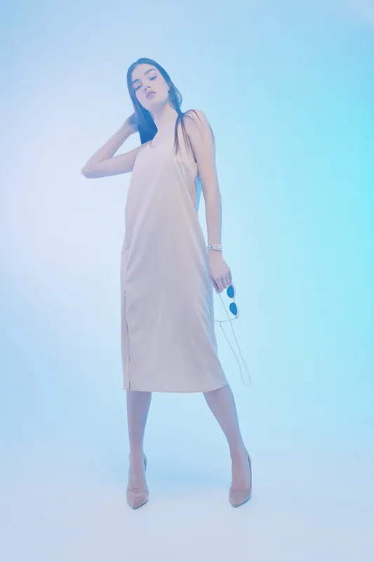 Woman beauty fashion model in a beige dress poses confidently holding sunglasses in a soft blue studio light. Concept for style, elegance, and modern clothing.