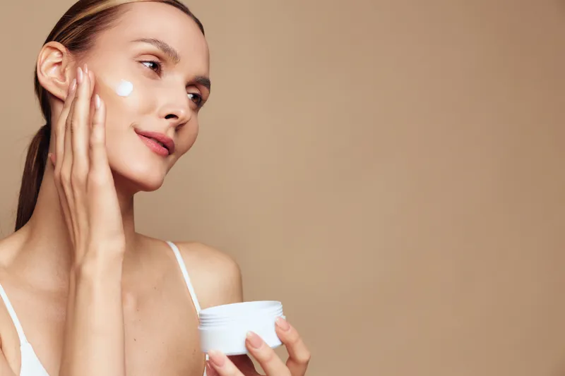 Young woman applies face cream on cheek holds jar with moisturizer has clean skin natural beauty and skincare routine isolated on beige background with positive expression