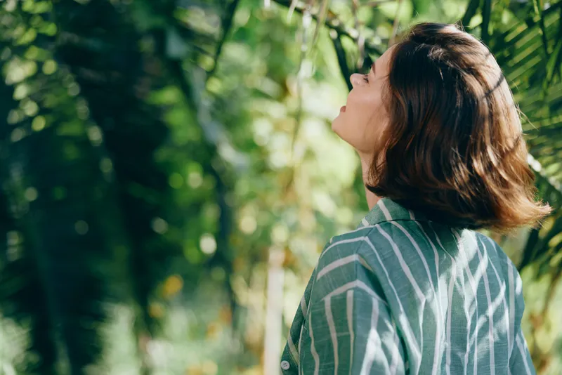 Portrait of a woman standing outdoors, looking up toward the sky as sunlight filters through lush green foliage.