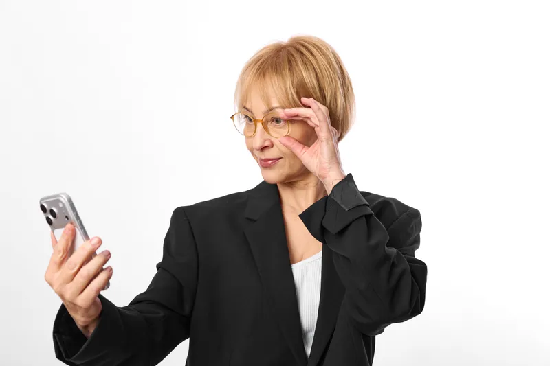 Senior woman businesswoman holding smartphone, adjusting glasses with one hand, surprised expression, looking at screen, focused on message, isolated on clean white background