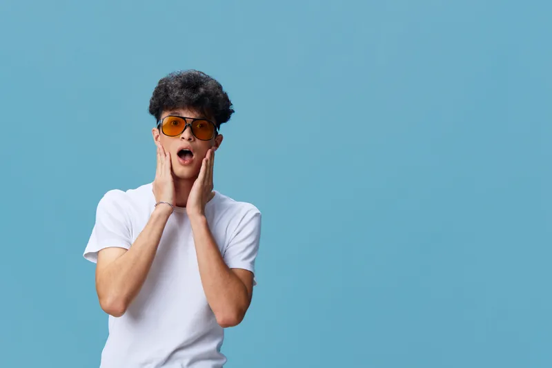 Surprised young man wearing sunglasses and white t-shirt with curly hair expressing shock against blue background in casual style.