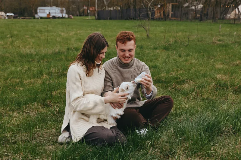 Smiling couple sitting on grass with a goat kid, outdoor lifestyle moment, warm natural light, gentle bonding, connection with animals, serene countryside mood.