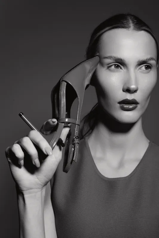 A young white woman with pale skin and slicked back hair holds a high heel shoe and a pen near her face in a black and white studio portrait. Concept of fashion, style, and creativity.