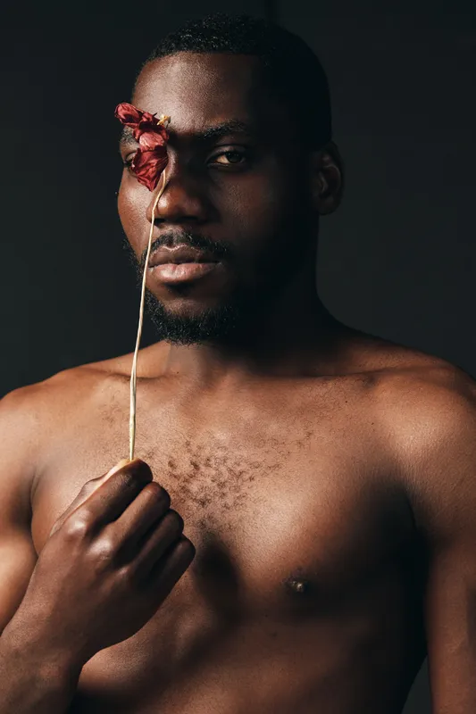 Creative conceptual portrait of African man holding dried flower in front of face with dark background, artistic visual style, bare chest, and thoughtful expression in studio lighting.