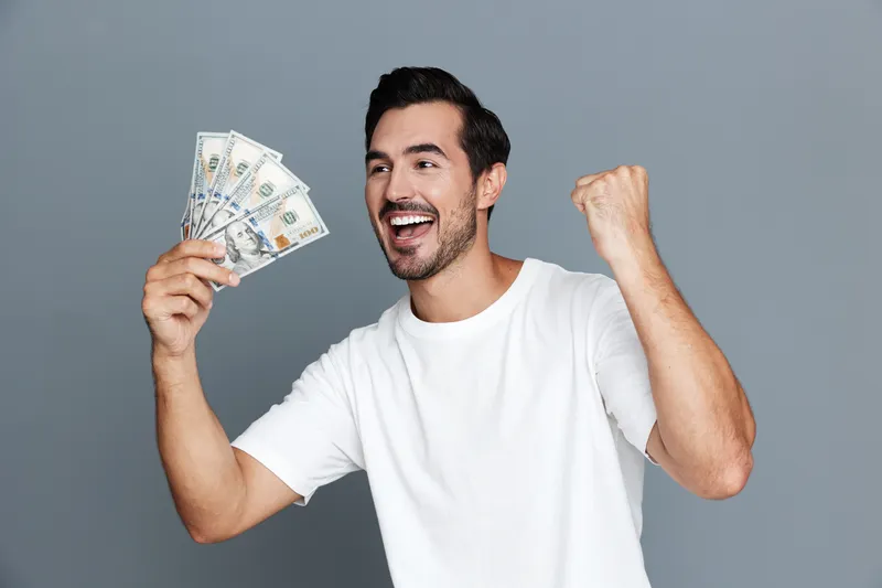 Happy cheerful young man celebrating success while holding dollar bills, showing excitement and joy in a minimalist gray background. People lifestyle concept