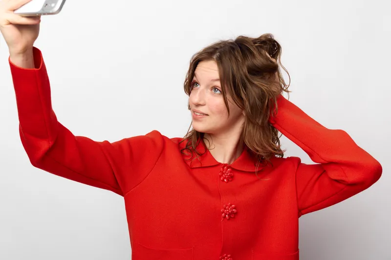 Young woman in red sweater takes a selfie with smartphone, looking away and touching hair. Casual style, isolated on white background, concept of social media and self-portrait.