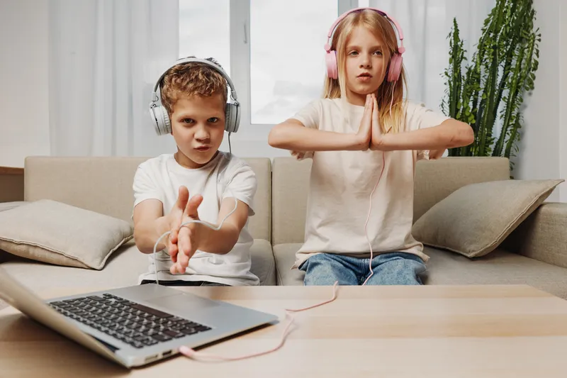 Two children with headphones sit on couch in living room using laptop, boy claps hands and girl meditates with closed eyes, casual home environment.
