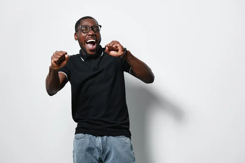 Excited black man with dark skin and glasses wearing black t shirt and jeans celebrating success with raised fists and open mouth on white background. Joyful african american male.