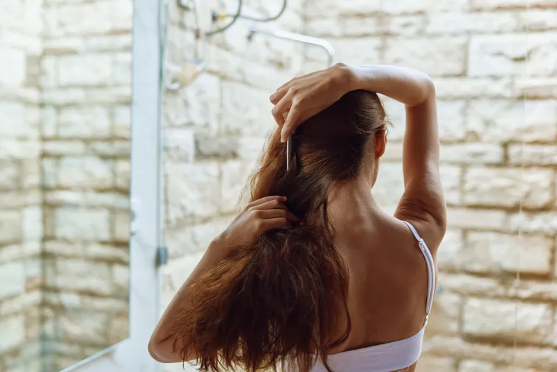 woman combing hair in bathroom with natural light, long brown hair, casual white top, minimalistic modern interior, morning routine