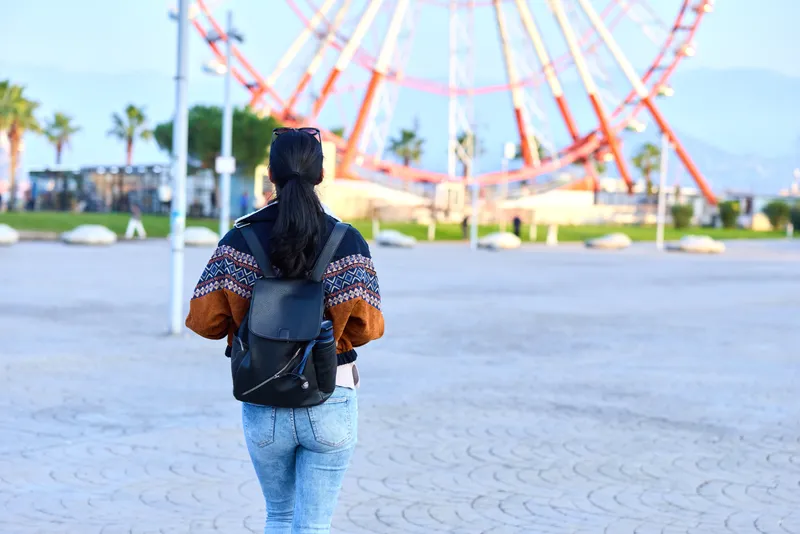 Woman with backpack stands on a boardwalk facing a colorful ferris wheel and palm trees, capturing a casual travel moment during a sunny seaside outing in the distance with a gentle breeze