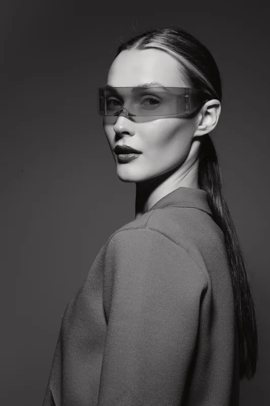 Portrait of a Caucasian woman with pale skin wearing transparent futuristic glasses and a textured jacket in a studio setting with dramatic lighting.