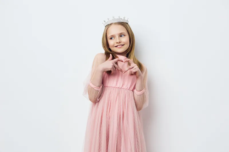 A young girl in a pink tulle dress and a crown, playfully poses with a charming smile against a neutral background, embodying innocence and joy