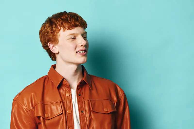 Redhead man with curly hair wearing brown leather jacket and white shirt posing looking aside on bright turquoise background. Young adult male portrait in casual style with confident expression.