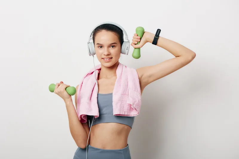 Young woman fitness portrait lifting green dumbbells while wearing headphones and a pink towel over shoulders in a minimalist studio setting, athletic outfit and confident workout expression.