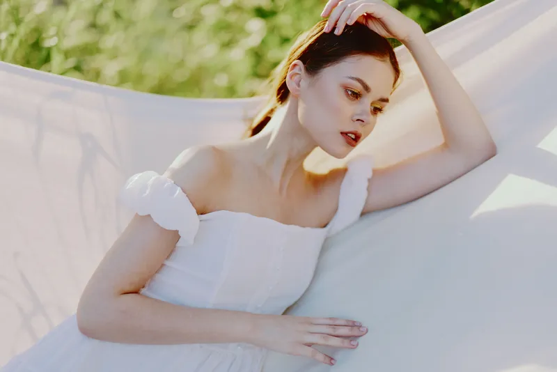 Young woman in a white dress with soft sleeves, posing gracefully outdoors, surrounded by greenery, exuding tranquility and elegance in a dreamy atmosphere