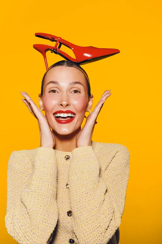 Happy woman with red high heel shoe on head smiles with hands near face on bright yellow background. Concept for fashion, fun, and playful lifestyle portrait.