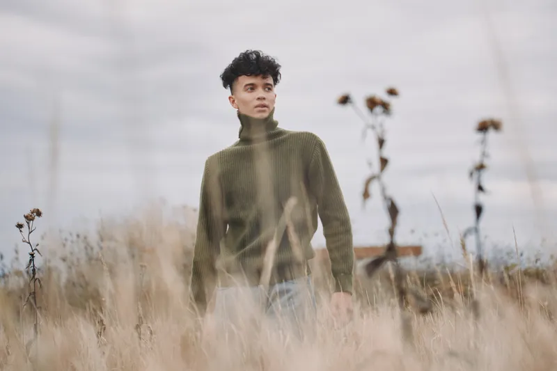 A young man stands in a muted field wearing a turtleneck sweater, captured in a candid, authentic moment that emphasizes genuine presence, natural expression and subtle emotion.