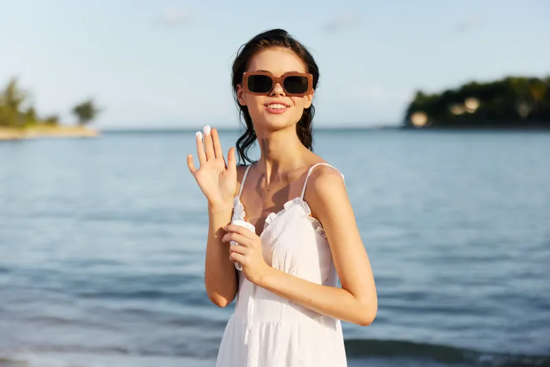 Smiling woman in white dress waving hand by the sea in summer sunny day with sunglasses and relaxed mood.