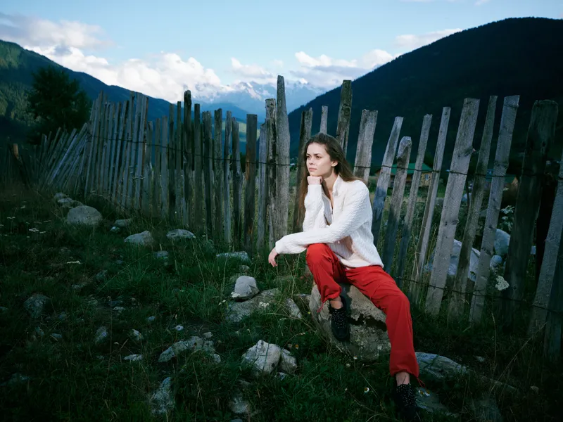Woman sits near a weathered wooden fence in an outdoor mountain landscape, wearing redpants and a white top, casual, tranquil mood in rugged terrain with rocky foreground