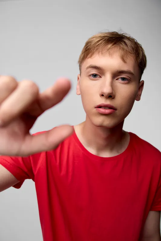 Young man in a red shirt extends his hand toward the camera in a close up studio shot, creating immediacy and connection with soft lighting and a neutral background