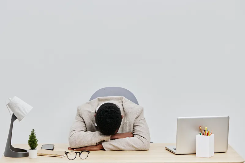 Modern black man with curly hair wearing beige sweater, bowing his head in frustration at cluttered desk with white background, office workspace, freelance professional, stress concept, flat lay, mini