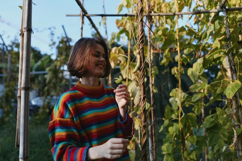 Woman in a rainbow striped sweater smiling outdoors in a backyard garden, short hair bob casually leaning on a vine trellis, relaxed lifestyle portrait in warm afternoon light