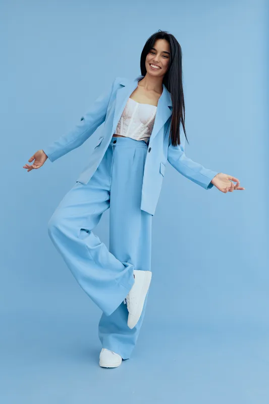 Happy woman in a stylish light blue suit and white sneakers poses joyfully against a solid blue background, showcasing modern fashion and youthful energy.