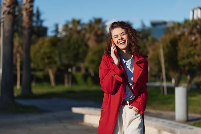 Young woman in a bright red coat chats on her phone outdoors, smiling in a sunny park as she strolls along a paved path, with a casual white shirt peeking out and trees framing the background.