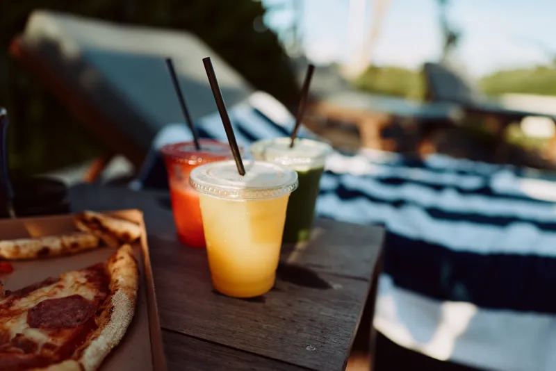 Outdoor scene with pizza on a wooden table, colorful drinks with straws, sunlit backyard, relaxed mood, casual dining, vibrant colors, leisure atmosphere and warm breeze