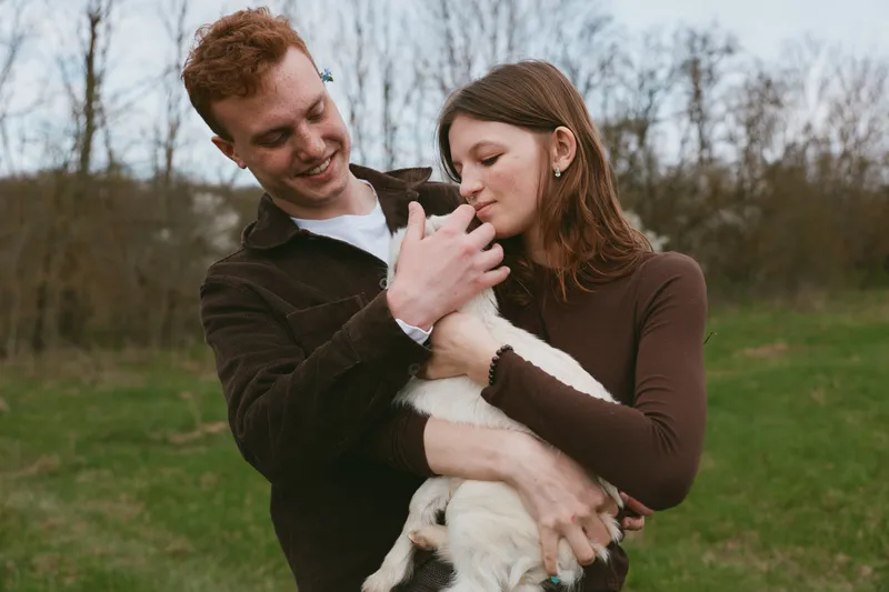 A couple outdoors bonds with a gentle goat kid, cradling the animal in a grassy field. Realistic lifestyle moment highlighting affection, animals, and family connection in a natural setting.