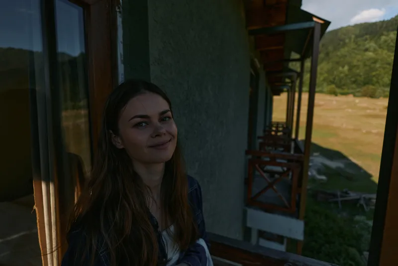 A cheerful young woman stands on a sunlit porch overlooking a green hillside, smiling softly and enjoying a calm outdoor moment surrounded by natural scenery, fresh air, and tranquil daylight.
