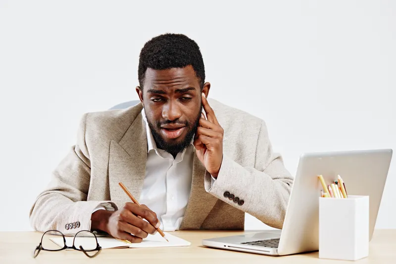Professional young African-American man in beige suit with confused expression, holding pen and talking on phone, sitting at desk with laptop and notebooks, isolated on white background studio portrai