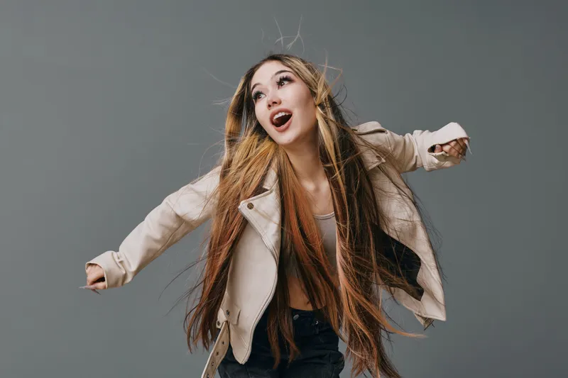 Young woman with long hair wearing beige jacket and black pants posing with arms outstretched against grey background in studio portrait of happy emotional person.