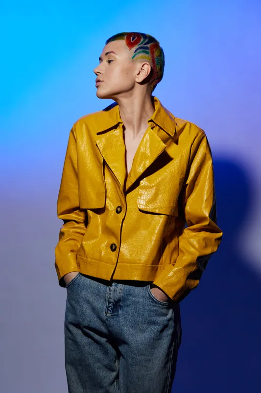 Young woman with colorful short hair wearing a yellow jacket and jeans, standing confidently against a vibrant blue and purple gradient background.