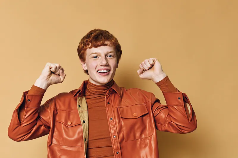 Young man with red hair wearing orange shirt and jacket smiling and posing with raised fists on beige background for positive emotion and success concept.