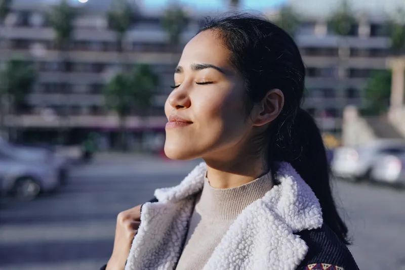Woman in a warm coat enjoys outdoor sunlight with serenity and calm, eyes closed as breeze brushes her hair, urban backdrop softly blurred, natural mood and relaxed ambiance