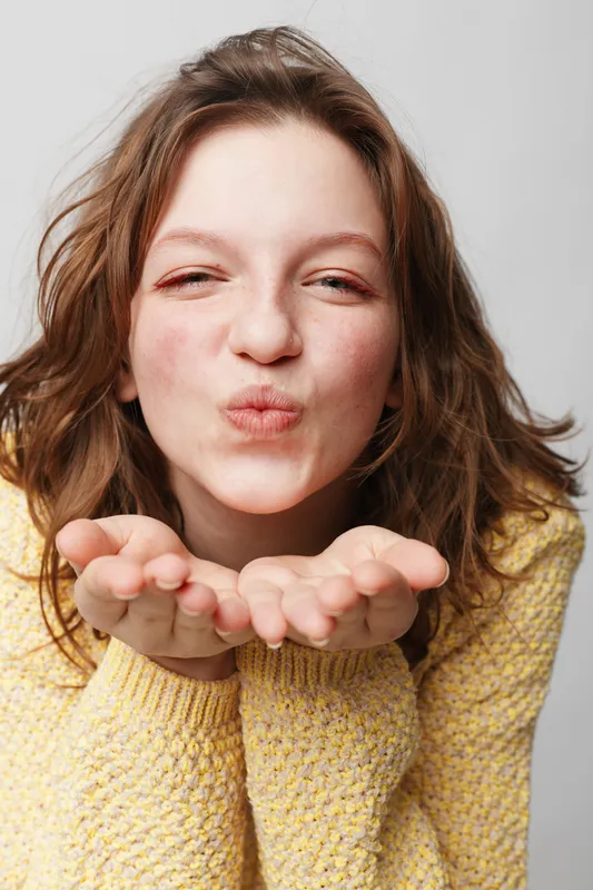 Young woman with wavy hair wearing yellow sweater blowing a kiss with closed eyes. Studio portrait on white background. Concept of affection, friendliness, and positive emotions.