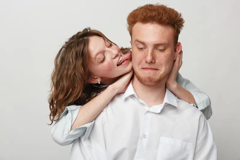 Playful young woman with curly hair teasing shy redhead man in white shirt on light background. Concept of fun, friendship, and casual interaction between young adults.