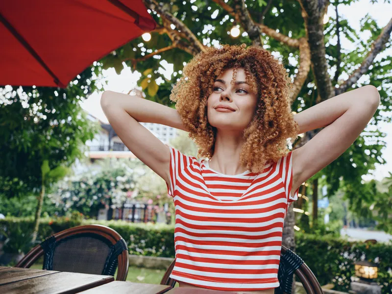 Young woman with curly hair wearing a red and white striped top lounges outdoors, arms raised behind her head, a relaxed smile, embodying a sunny casual summer mood.