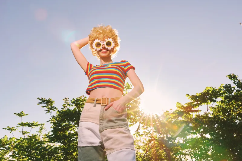 Young woman outdoors in a striped top and high waisted shorts smiles behind flower sunglasses as sunlight pours over a carefree colorful summer moment
