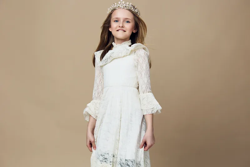 A joyful young girl wearing a white dress and a crown, standing against a neutral background, radiating happiness and innocence, perfect for children s fashion concepts
