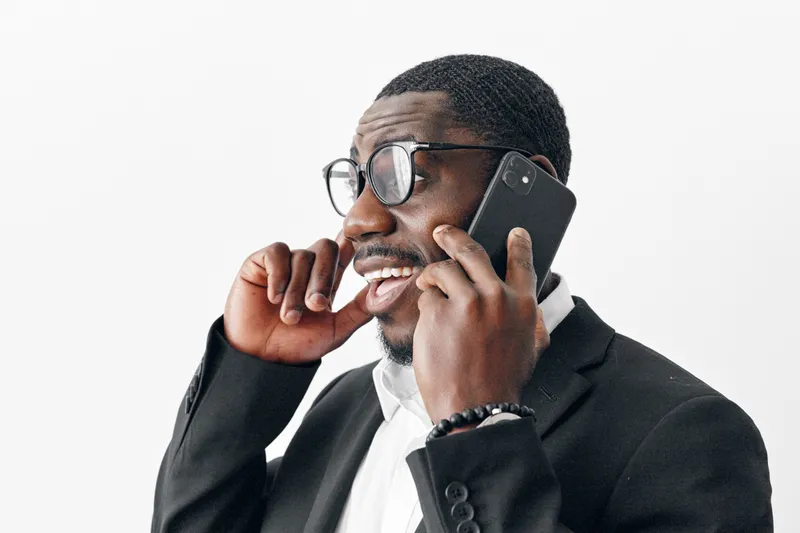 African American man with dark skin wearing glasses and black suit talking on smartphone and smiling with finger near mouth on white background.