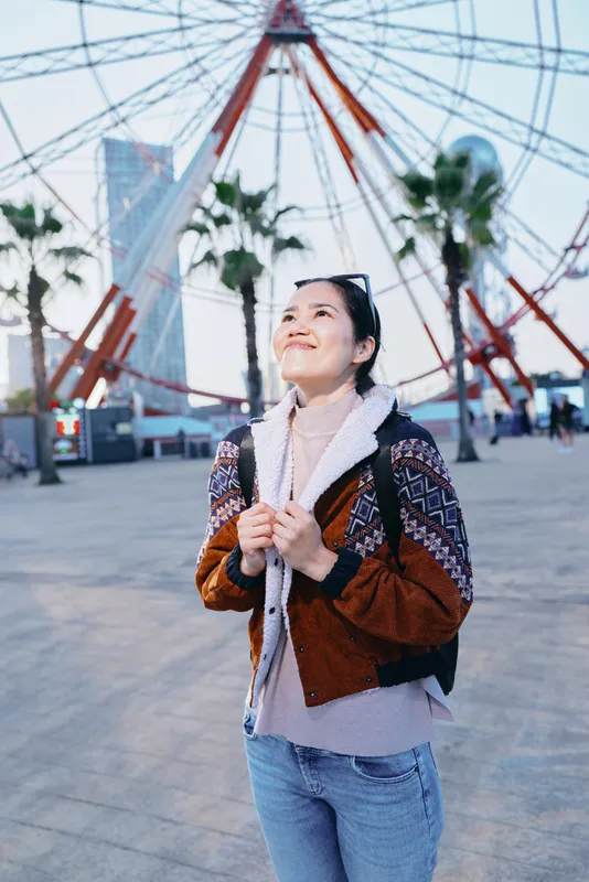Woman in brown jacket and scarf stands outdoors in an amusement setting, gazing up near a large ferris wheel, palm trees and city buildings in the backdrop, sunny, relaxed mood and candid travel vibe